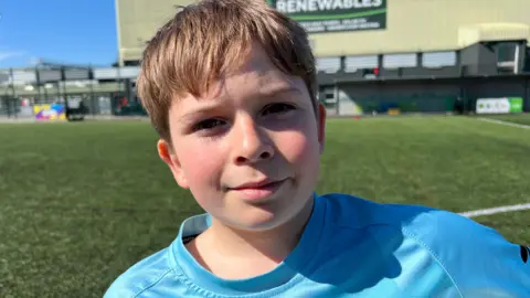 A boy wearing a blue football top. He has brown hair and is smiling. Behind him is the LNER Stadium.