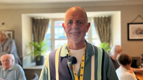 Ian is wearing stripped shirt with different shades of blue and a yellow lanyard. He has a short grey beard and cropped white hair. He is smiling at the camera.