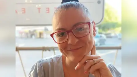 Family handout A woman with dark brown pulled back hair is wearing red glasses and smiling at the camera, she has one hand up to her face and a diamond ring is visible on her fourth finger. She's wearing an off-white top and appears to be sitting outdoors as a blurry car park is visible behind her