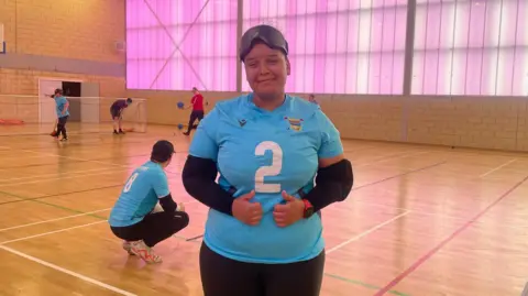 Goalball player Alex Ulysses is wearing a blue sports top with the number two in white on the front of it. She is stood in a sports hall with other players in the background. There are a set of goals and a wall which is illuminated with purple light.