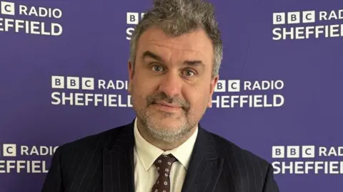 A middle-aged man, with grey hair and beard, wears a pin-stripe black suit with a brown tie. He is standing against a purple BBC Radio Sheffield backdrop.