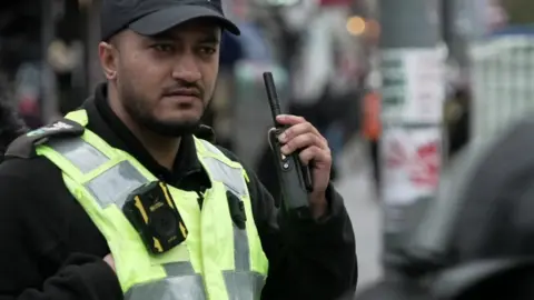 A male member of the drug squad, wearing a hi-vis jacket with a body worn camera on the front. He has a baseball cap on his head. He is on a street, with a radio raised in his hand