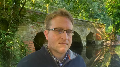 Matt Marvel/BBC A man with short hair wearing glasses and a blue sweater standing in front of a bridge