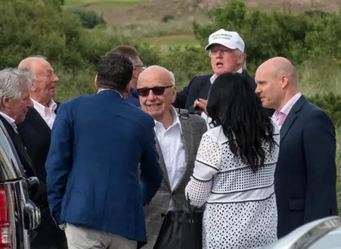 Pa Trump i Murdoch są w tłumie ludzi na międzynarodowym polu Golf Links. Trump jest z tyłu w białej czapce Maga, Murdocha przed nim, wydając się uścisnąć czyjąś rękę. Murdoch ma na sobie białą koszulę i szarą marynarkę.