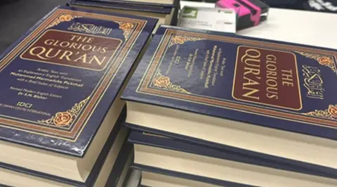 Razia Hadait More than six Qurans, with a mainly blue cover, are horizontal. Three are piled up on the left, with three on the right. The words The Glorious Qur'an are in large letters on the front.