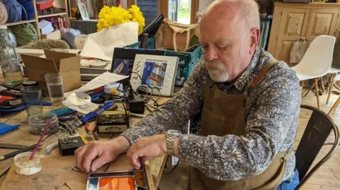 Roland Berry, pictured in his workshop. He is working with a panel of stained glass and is wearing a floral shirt and a brown apron. The workshop has lots of tools and books in the background, as well as a vase of daffodils and shelves of large balls of yarn. 