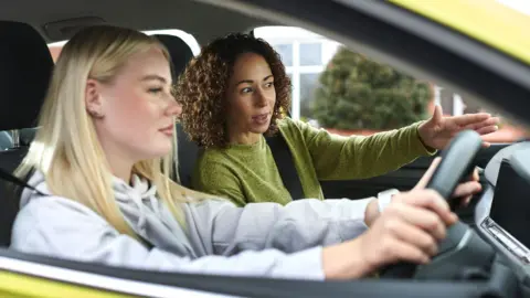 A young woman sitting behind the wheel of a car about to take her driving test. A female driving test examiner is speaking to her first.