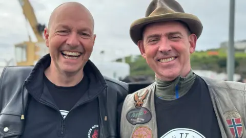 Ole Skaar Erling and Thomas Pettersen standing next to each other with smiling. Ole is bald and is wearing a black leather coat and black hooded jumper. Erling is in a black top and green waistcoat covered with badges and a camel-coloured hat.



