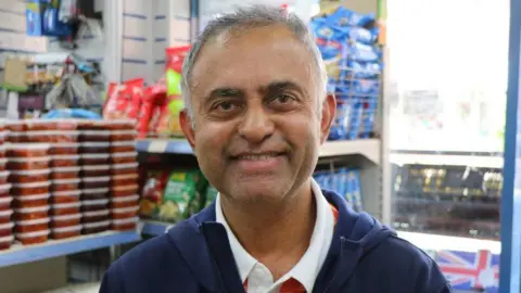 Dharmesh Patel, a middle-aged Indian man, standing inside a grocery shop in Leicester. He is wearing a navy hoody over a polo shirt.