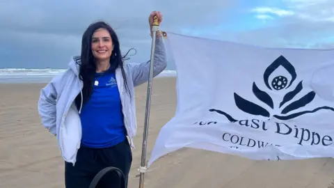 Ann-Marie Harkin Ann-Marie Harkin has long dark brown hair is standing on the beach beside a white flag which is blowing in the wind. It has a black logo which looks like a flower and the words Seacoast Dippers in black type. She is wearing a bright blue t-shirt under a gray zip up hoody and a pair of dark coloured tracksuit bottoms. Behind her you can see long swathes of sand and white-tipped waves. 