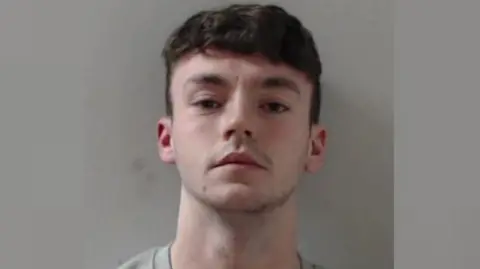 A man with dark hair looks at the camera with an expressionless face for a police mugshot