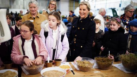 Duchess of Edinburgh looking at bowls of maize and grass, alongside schoolchildren