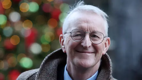 EPA A close-up of Hilary Benn standing outside Downing Street after a cabinet meeting. The image is a head and shoulders shot of Benn, who is smiling. He is an older man, with short, white hair and he is wearing a pair of circular metal glasses, a tweed coat and a blue collared shirt. He is standing in front of a lit up Christmas tree, but the lights are blurred.