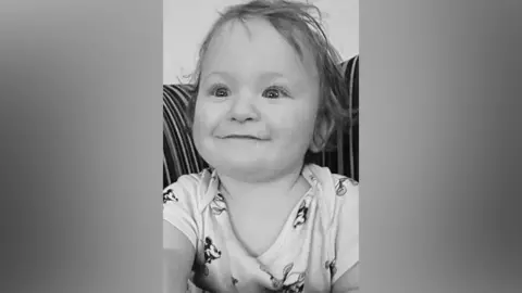 A black and white picture of a smiling Louie Hughes. The baby is wearing a Mickey Mouse vest.