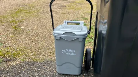 Vikki Irwin/BBC A small grey caddy is on the ground next to a large black wheelie bin. The grey bin has a large black handle and says "Suffolk recycles" on it. 