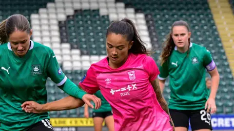 Ipswich Town Football Club Natasha Thomas is pictured during a football game. She is attempting to tackle a ball away from a player on the opposing team. She is looking down while attempting to avoid the other player. She is wearing the pink Ipswich Town away kit. Another player can be seen in the background as well as the stands of the football ground.