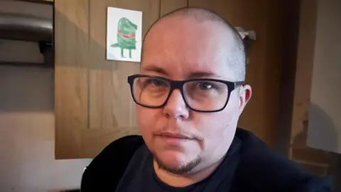 Vince is white and wears black-framed glasses. He has a shaved head and a brown goatee with a slight moustache. He is standing in his kitchen and looking into the camera's lens. There are brown kitchen cabinets behind him, with a child's drawing displayed on one of them.