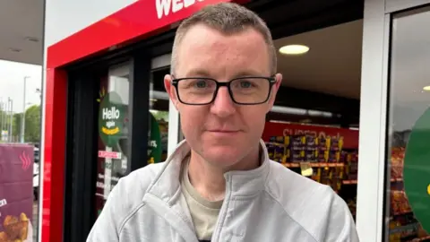A man with short brown hair looks into the camera. He is wearing black squared glasses and a light grey pullover with the word 'O'Neill's' stitched on. He is standing at the doorway of a shop.