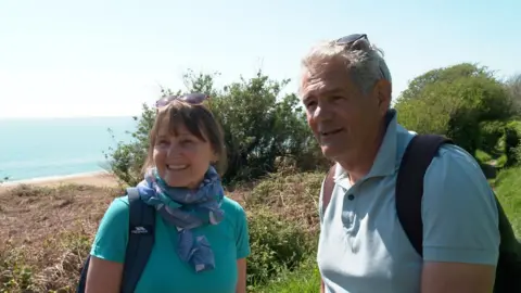 A woman with sunglasses on her head wearing a scarf smiles at the camera. The sea at Strete Gate and the South West Coast Path are behind her. She is joined by her husband wearing a blue top and carrying a backpack.
