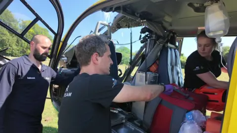 A bald, white middle-aged man with a beard wearing a dark long-sleeved top standing next to a white middle-aged man with dark-greying hair wearing a t-shirt, both looking in to a helicopter that is stationary in a field, with a young white woman in black t-shirt and orange trousers who is sitting inside