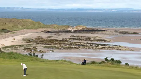 Getty Images Matt Fitzpatrick of England putts on the 13th green during the pro-am prior with the beach and the North Sea in the background. He is wearing a white cap and jumper, grey trousers and white golf shoes. He is a small figure in the bottom left of the picture, which focuses on the background scenery