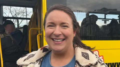 BBC/Abi Jaiyeola Georgie Grove stands in front of a yellow bus. She has tied back brown hair and is wearing a white fleece and denim top.