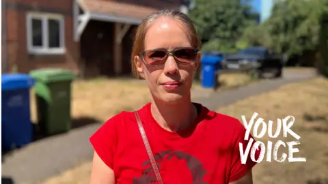 A young woman with blonde hair tied back in a red t-shirt and sunglasses stands outside a house. There is yellowed grass in the background of the image. 