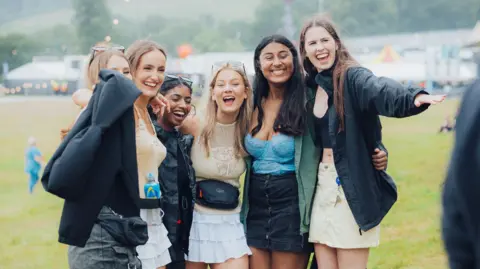 Jessie Myers A group of six young women, dressed in summery outfits with jackets on top, smiling as they pose arm in arm.