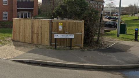 A street sign reading Southfields Way. It stands in front of a light brown wooden fence. There is a pavement in front of it.