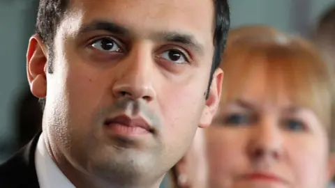 PA Media Anas Sarwar with a stern expression looking into the distance. He has black hair and looks to be wearing a suit. A woman with blonde hair is blurred in the background.