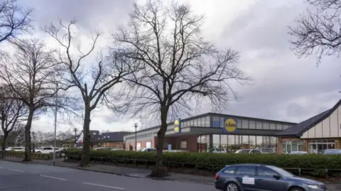 A computer generated image of the Lidl store next to retained units at Fallowfield Retail Park, with cars parked in front of the store, fronted by trees and bushes and a car is shown driving along the road in front of the site