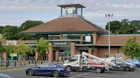 Google Streetview of several cars parked in a car park with a large supermarket behind. It has the sign saying Morrisons above large glass doors and has a little tower surrounded by brown slanted roofs.