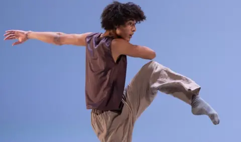 Jakob dancing, on a stage, with a blue background. He is wearing a brown top, brown trousers and grey socks. His left arm is outstretched and right arm across his body. His leg is raised. He has short curly dark hair. 