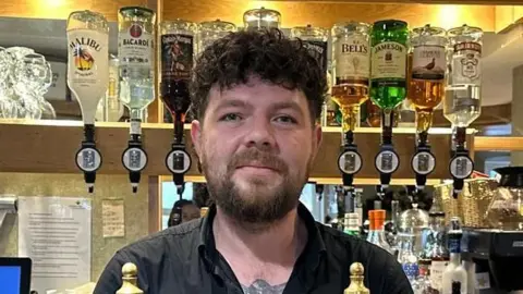Jay Kemsley is standing behind the counter at a bar. He has short brown, curly hair and and a beard. Various spirits are hung up on the wall behind him.
