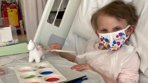 Family photograph Chloe with short brown hair lies in a hospital bed with a face mask on. She is painting a ceramic pony with a paint pallet filled with coloured paint and brushes on her tray. 