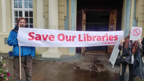 Campaigners outside the library protesting against the decision to re-locate the service. Two campaigners are holding a sign which says "Save our Libraries".