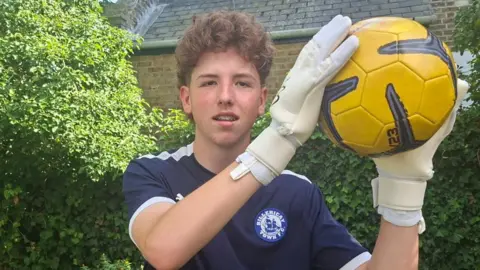 Freddy stands outside, with a green bush behind him. He holds a yellow football aloft, wearing white goalkeeping gloves. He wears a navy blue shirt with a blue crest, which is printed with a drawing of a sailing ship and the words BILLERICAY TOWN FC.