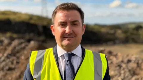 Colin O'Hanlon. He has short dark hair, wearing a yellow high viz vest with navy blazer, white shirt and navy tie underneath. He is standing on wasteland with muck hills in the background. 