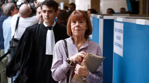 EPA Gisèle Pelicot, a woman with a red bob and a purple blouse carrying a folder of documents, is walking through a room, followed by Stéphane Babonneau, a man with dark hair in a formal black gown