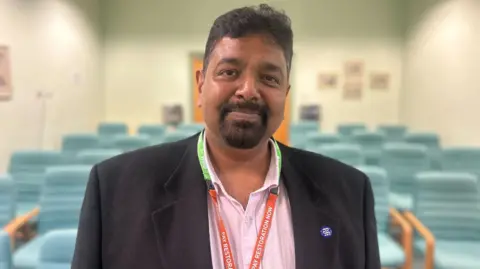 Prakash Thiagarajan smiling. He is wearing a dark suit jacket and pink shirt and standing in front of some teal green chairs.