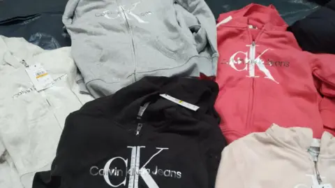 A selection of hoodies are spread out on a table. Two are light coloured, others are blue, grey and pink. all have Calvin Klein Jeans on them.
