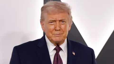 Donald Trump in a blue suit and blue tie with red dots stands in front of a white background