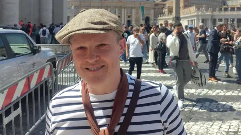 Ronan Mac Murchaidh is standing in the middle of a busy square in the Vatican City and is smiling at the camera. He is wearing a flat cap, as well as a skinny brown scarf and a white and black striped top.