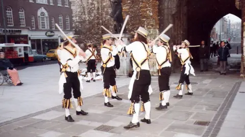 Five Morris men from King John's Morris Men wearing top hats white shirts, black trousers and white socks pulled up to the knee. They have yellow and black tassels and accessories. They are dancing in front of the bargate in Southampton at dawn 