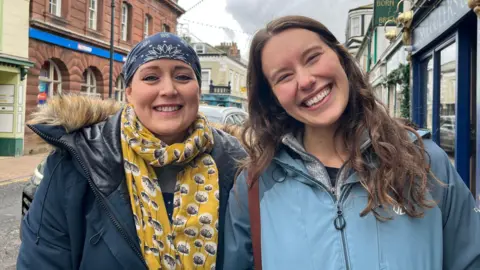 Federica Bedendo/BBC Gemma Bellamy and Monica Wilson standing outside shops in Maryport. Ms Bellamy is wearing a blue headscarf, yellow scarf and blue coat. She has blue eyes and is smiling at the camera. Ms Wilson has long, brown, wavy hair, is wearing a light blue coat and is also smiling.