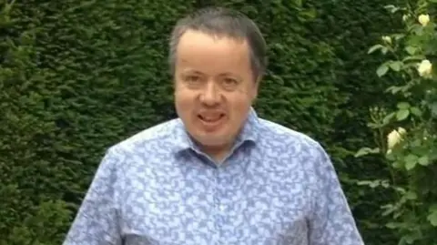 Family photo A man with grey hair smiling at the camera in front of green bushes. He is wearing a short sleeved blue patterned shirt.
