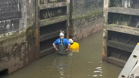 Two people stand in shallow water inside a canal lock wearing white safety helmets. One is in a blue T-shirt and chest waders, the other in a yellow high-visibility jacket. The lock's wooden gates and blackened brick walls rise steeply on either side.