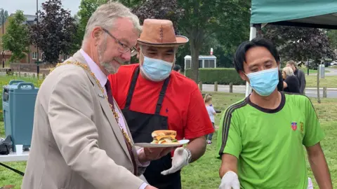 Nor Aziz A man with short grey hair and a grey beard and glasses stands holding a plate of food. He is wearing a light brown or grey suit over a pink pinstripe shirt and a burgundy tie. Two other men are nearby wearing facemasks. One has black hair in a centre parting and is wearing a green t-shirt. The other is wearing a black apron over a red t-shirt, and a hat.