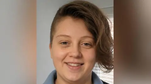 Family photo Menna, a young woman with short dark hair which is shaven at the sides, and has a long quiff at the front. It is a head and shoulders shot of her, with a window with blinds behind her. 