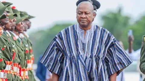 President John Dramani Mahama wearing a fugu attire with blue and white strips. He is inspecting a guard of honour. 